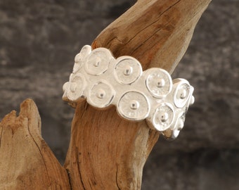 May include: A silver ring with a textured design of overlapping circles. The ring is displayed on a piece of driftwood.