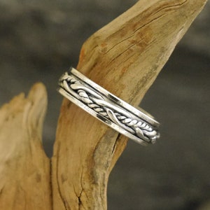 Könnte beinhalten: Ein silberner Ring mit einem geflochtenen Design. Der Ring liegt auf einem Stück Treibholz.