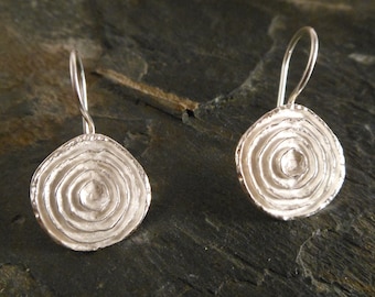 May include: A pair of silver earrings with a circular design featuring concentric circles. The earrings have a simple, elegant style.