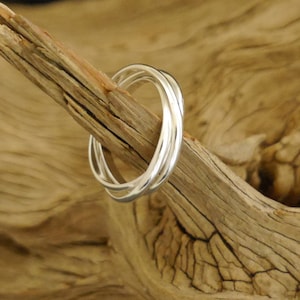Könnte beinhalten: Ein silberner Ring mit drei ineinandergreifenden Bändern. Der Ring liegt auf einem Stück Treibholz.