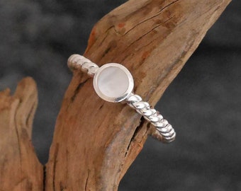 May include: A silver ring with a twisted band and a round, white mother-of-pearl stone. The ring is displayed on a piece of driftwood against a dark background. The ring's band has a rope-like design.