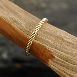 Könnte beinhalten: Ein goldfarbener Ring mit einem gedrehten Seildesign. Der Ring ist auf einem Stück hellbraunem Holz mit sichtbarer Maserung platziert. Der Hintergrund ist eine dunkle, verschwommene Oberfläche.