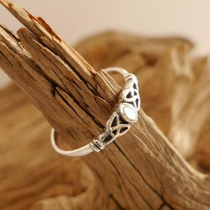 Könnte beinhalten: Ein silberner Ring mit einem keltischen Knotenmuster und einem kleinen, oval geformten weißen Stein. Der Ring ist vor einem Hintergrund aus strukturiertem, hellbraunem Holz platziert.