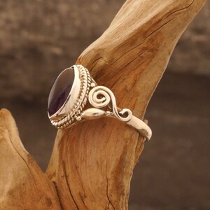 May include: A silver ring with a purple gemstone set in a bezel setting. The ring has a decorative swirl design on the band.