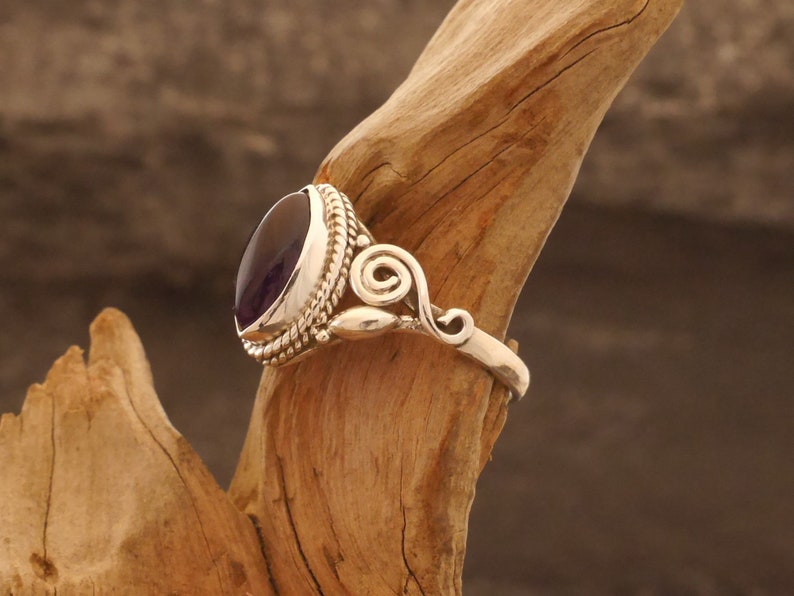 May include: A silver ring with a purple gemstone set in a bezel setting. The ring has a decorative swirl design on the band.