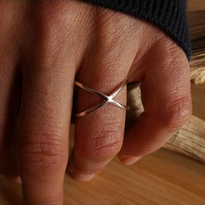 Könnte beinhalten: Ein silberfarbener Ring mit einem X-förmigen Design. Der Ring, der an einem Finger getragen wird, besteht aus zwei sich kreuzenden Bändern. Er hat eine schlichte, moderne Ästhetik mit poliertem Finish. Ein modernes Schmuckstück.
