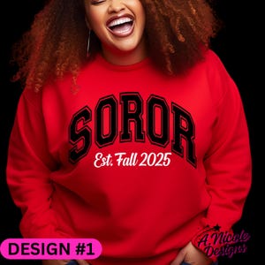 May include: A red sweatshirt with the word "SOROR" in black block letters outlined in white. Below the word "SOROR" is the text "Est. Fall 2025" in white. The sweatshirt also has the text "DESIGN #1" in a pink rectangle.