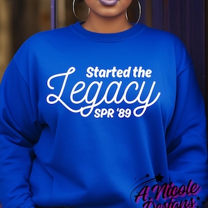 May include: Royal blue sweatshirt with the white text "Started the Legacy SPR '89". The design includes a cursive "Legacy" and the text "A Nicole Designs" in the bottom right corner. The sweatshirt is a solid color.