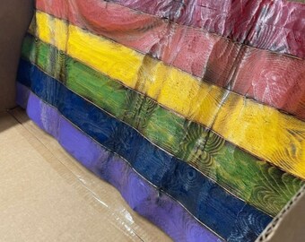 May include: A rainbow pride flag made of wood planks. The flag is folded and placed inside a cardboard box.