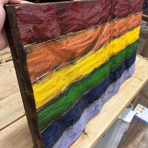 May include: A wooden wall art piece featuring a rainbow flag design. The artwork is made of stained wood planks, with each plank painted a different color of the rainbow. The edges of the wood are dark brown, and the colors are vibrant.