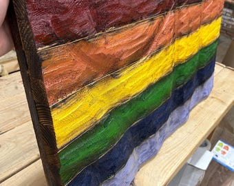 May include: A wooden wall art piece featuring a rainbow flag design. The artwork is made of stained wood planks, with each plank painted a different color of the rainbow. The edges of the wood are dark brown, and the colors are vibrant.