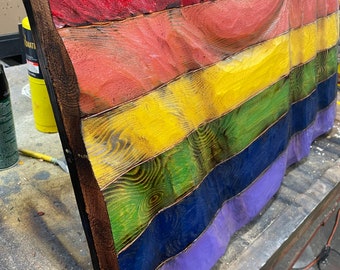 May include: A wooden rainbow flag with a textured surface. The flag is painted in the colors of the rainbow, with red, orange, yellow, green, blue, and purple stripes. The flag is mounted on a wooden frame.