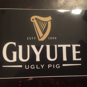 Puede incluir: Logotipo en blanco y negro sobre fondo negro con un diseño de arpa dorada. El logotipo dice "Guyute Ugly Pig" y "Est. 1994".