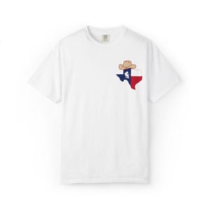 May include: White t-shirt featuring a Texas state outline in red, white, and blue, with a cowboy hat and a silhouette of a person. The shirt is made of a soft material and has a classic crew neck.