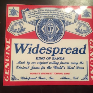 Peut inclure: Un autocollant rouge, blanc et bleu avec le texte "GENUINE" sur les côtés gauche et droit. L'autocollant présente un logo avec le texte "Widespread" et "KING OF BANDS". Le texte "Made by our original writing process using the Choicest Jams for the World's Best Fans" est également inclus. L'autocollant comprend également le texte "Widespread Panic, Inc. Athens, GA".