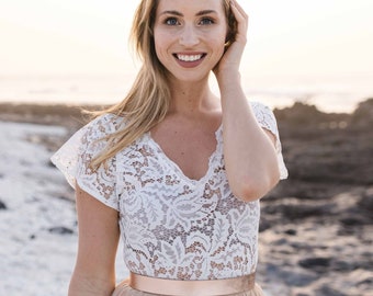 May include: A woman wearing a white lace top with a rose gold belt and a beige tulle skirt. She is standing on a beach with a sandy background.