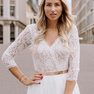 May include: A woman wearing a white lace top with a gold belt and a white tulle skirt. She has long blonde hair and is wearing gold earrings.