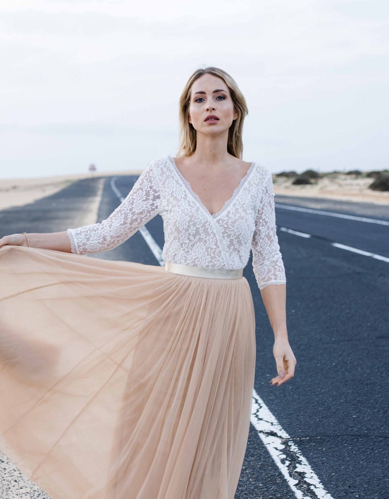 May include: A woman wearing a white lace top with a satin belt and a long, flowing beige tulle skirt. She is standing on a paved road with a desert landscape in the background.