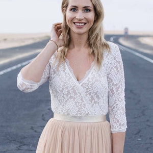 May include: A woman wearing a white lace top with a v-neckline and a long, flowy blush-colored tulle skirt. She is standing on a paved road with a sandy background.