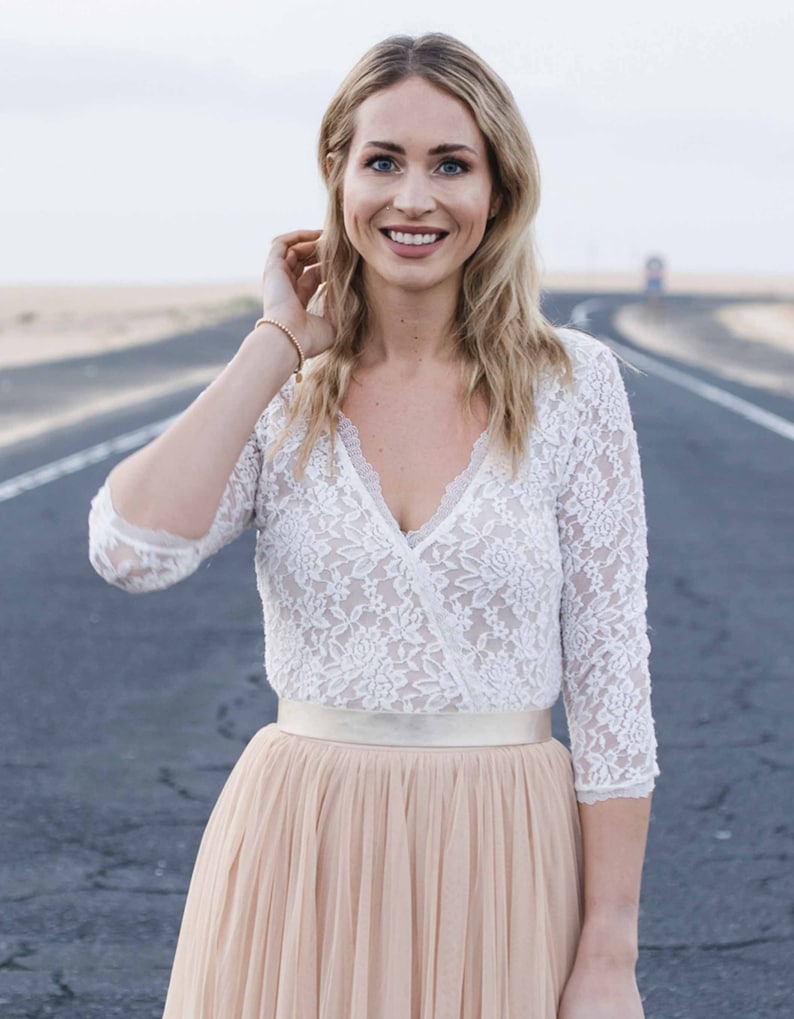 May include: A woman wearing a white lace top with a v-neckline and a long, flowy blush-colored tulle skirt. She is standing on a paved road with a sandy background.