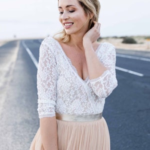 May include: A woman wearing a white lace top with a plunging neckline and a long, flowing beige skirt. She is standing on a paved road with a desert landscape in the background.