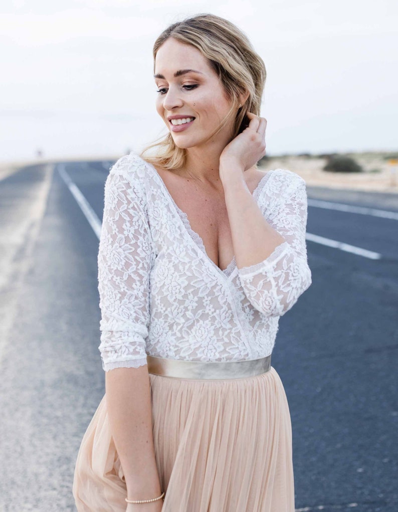 May include: A woman wearing a white lace top with a plunging neckline and a long, flowing beige skirt. She is standing on a paved road with a desert landscape in the background.