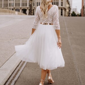 May include: A white tulle skirt with a lace top and a gold belt. The skirt is flowing and the woman is walking on a paved street.