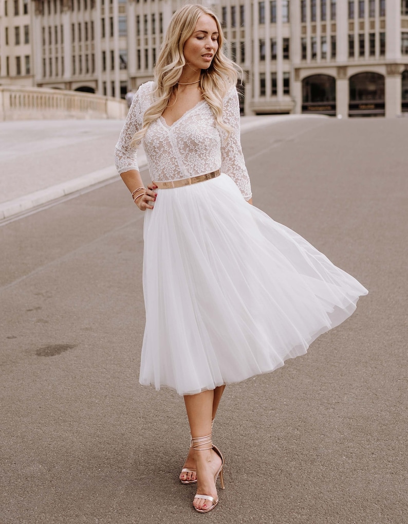 May include: A white lace top with a v-neckline and 3/4 sleeves paired with a white tulle skirt and a gold belt. The woman is wearing gold heels.