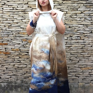 Puede incluir: Un vestido largo, blanco, con capucha y un diseño abstracto marrón y azul. El vestido tiene un cinturón y está hecho de una tela suave y fluida.