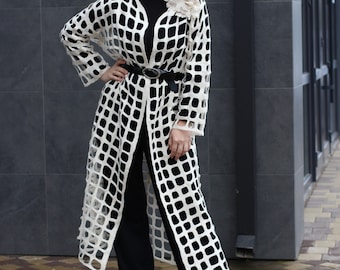 Può includere: Un cardigan bianco a trama aperta con una cintura nera e una spilla a forma di fiore bianco. Il cardigan ha una silhouette lunga e fluida e uno schema quadrato.
