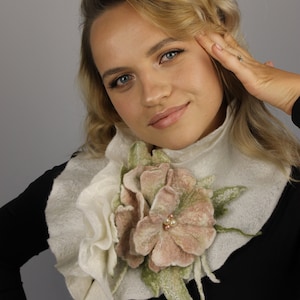 May include: A white felted flower collar with a pink flower and green leaves. The collar is worn by a person with blonde hair.