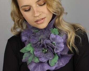 May include: A purple and green felt flower neck warmer with a green gem in the center. The neck warmer is worn by a person with long blonde hair.