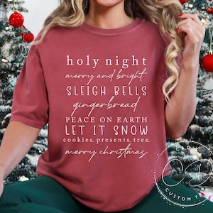 May include: A woman wearing a maroon t-shirt with white text that reads "holy night merry and bright sleigh bells gingerbread peace on earth let it snow cookies, presents, tree. merry christmas". She is sitting on a white and red striped rug with a gingerbread house and a Christmas tree in the background.