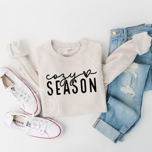 May include: A beige sweatshirt with the text "cozy season" and a small heart graphic. The sweatshirt is paired with blue jeans and white sneakers.