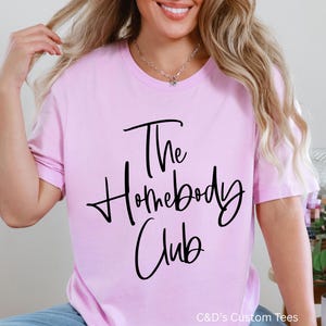 Puede incluir: Camiseta rosa claro con la frase "The Homebody Club" en escritura negra. La camiseta de manga corta se combina con vaqueros azules y zapatillas blancas. La imagen tambi&eacute;n muestra una bola de discoteca y plantas en macetas.