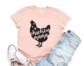 May include: A pink t-shirt with a black silhouette of a chicken with the text "chicken mama" inside the silhouette. The shirt is laid flat on a white background with a pair of white sneakers and a pair of light blue jeans.