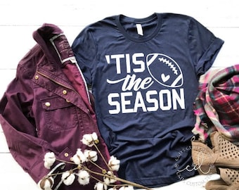 Könnte beinhalten: Ein marineblaues T-Shirt mit einer weißen Grafik, auf der "'Tis the season" mit einem Football und einem Herzen steht. Das Shirt liegt auf einer weißen Holzoberfläche mit einer burgunderfarbenen Jacke, einem karierten Schal und einem Paar braunen Cut-Out-Stiefeletten.