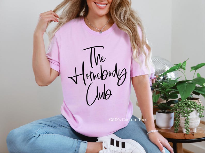 Puede incluir: Camiseta rosa claro con la frase "The Homebody Club" en escritura negra. La camiseta de manga corta se combina con vaqueros azules y zapatillas blancas. La imagen tambi&eacute;n muestra una bola de discoteca y plantas en macetas.