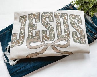 May include: Cream-colored t-shirt with the word "JESUS" in a camouflage pattern. A brown bow is centered below the text. The shirt is folded on top of a pair of blue denim jeans.