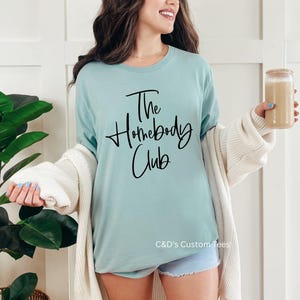 Puede incluir: Una camiseta azul claro con el texto "The Homebody Club" en escritura negra. La camiseta se combina con pantalones cortos de mezclilla y una rebeca color crema. Se sostiene un vaso de caf&eacute; helado en la mano.