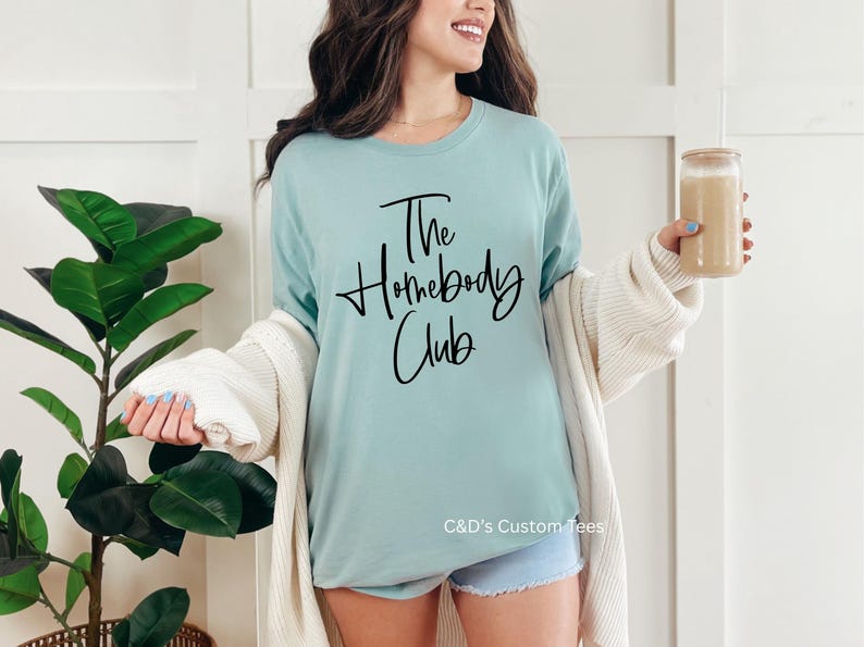 Puede incluir: Una camiseta azul claro con el texto "The Homebody Club" en escritura negra. La camiseta se combina con pantalones cortos de mezclilla y una rebeca color crema. Se sostiene un vaso de caf&eacute; helado en la mano.