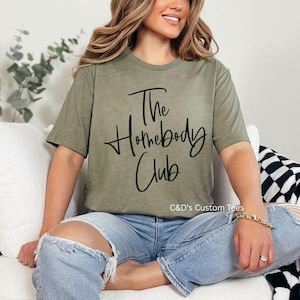 Puede incluir: Camiseta verde oliva con el texto "The Homebody Club" en escritura negra. La camiseta se usa con vaqueros azules. La imagen tambi&eacute;n muestra un sof&aacute; blanco y una manta a cuadros blancos y negros.