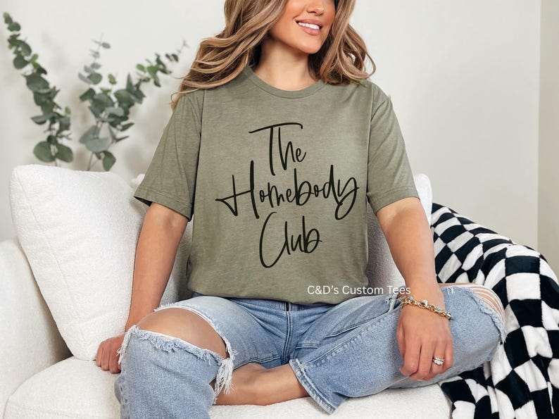 Puede incluir: Camiseta verde oliva con el texto "The Homebody Club" en escritura negra. La camiseta se usa con vaqueros azules. La imagen tambi&eacute;n muestra un sof&aacute; blanco y una manta a cuadros blancos y negros.