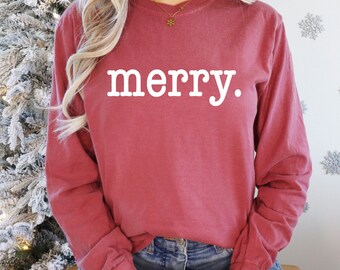 Könnte beinhalten: Eine Frau trägt ein langärmeliges, maroonfarbenes T-Shirt mit dem Wort "merry" in weiß auf der Vorderseite. Sie hält eine rot-weiß karierte Tasse mit Marshmallows.