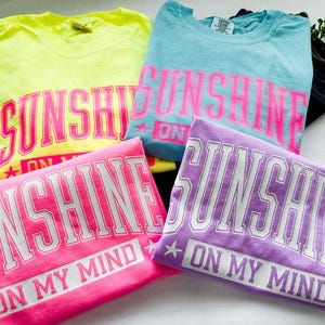 May include: Four folded t-shirts in bright colors: yellow, light blue, pink, and purple. Each shirt features the text "SUNSHINE ON MY MIND" in a contrasting color. The shirts are neatly stacked, showcasing the design.