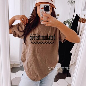 May include: A brown t-shirt with black text that reads "probably overstimulated so please don't talk to me". The shirt is being worn by a person with brown hair and a brown and white baseball cap. The text "C&D's Custom Tees" is visible at the bottom of the image.