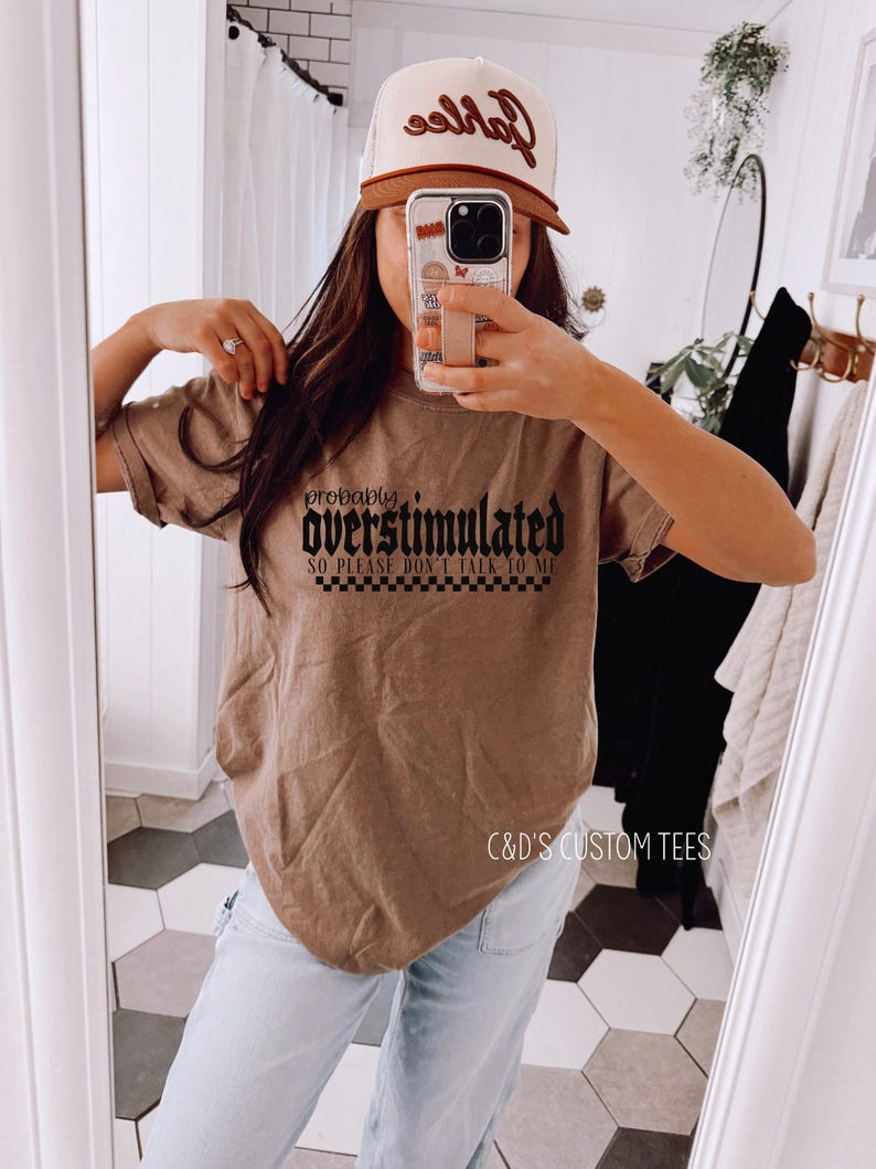 May include: A brown t-shirt with black text that reads "probably overstimulated so please don't talk to me". The shirt is being worn by a person with brown hair and a brown and white baseball cap. The text "C&D's Custom Tees" is visible at the bottom of the image.