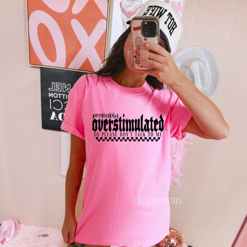 May include: Pink t-shirt with black text that reads "probably overstimulated so please don't talk to me" with a black and white checkered pattern underneath the text.