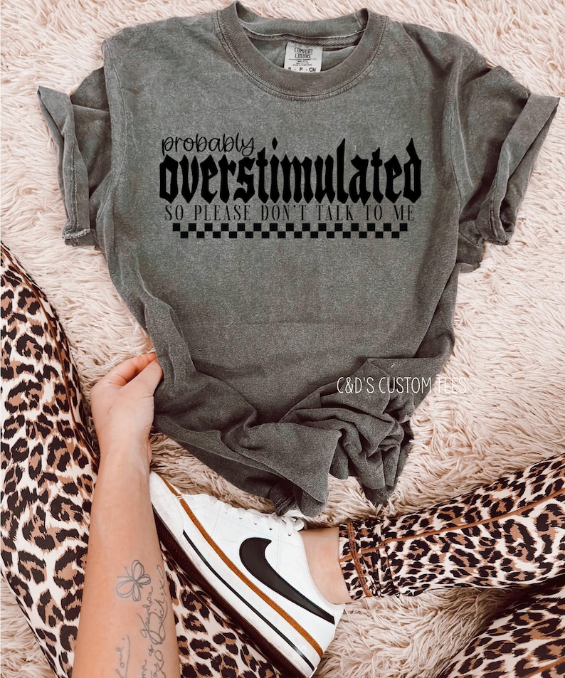May include: A gray t-shirt with the text "probably overstimulated so please don't talk to me" in black lettering with a checkered pattern underneath.