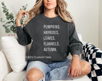May include: Dark gray sweatshirt with the words "PUMPKINS. HAYRIDES. LEAVES. FLANNELS. AUTUMN." in white block letters. The sweatshirt is part of the fall season theme. The brand name "C&D's Custom Tees" is printed at the bottom.
